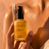 Hand holding a bottle of 'Comforting Face & Body Oil' by Coopers against a sunlit skin background.