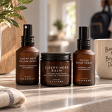 Three Coopers skincare products in amber glass bottles and a jar, arranged on a bathroom counter with a plant, mug, and towel nearby.